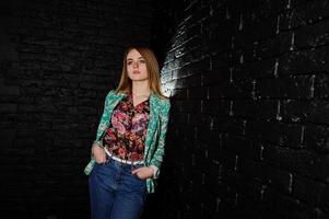 Stylish blonde girl in jacket and jeans against brick black wall at studio. photo