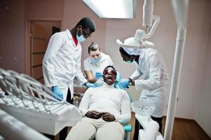 Multiracial dentist doctors team. African american man patient at UV protective glasses. His teeth treated with the help of a dental UV curing light lamp and a dental mirror. photo