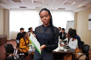 Face of handsome african business woman, holding clipboard on the background of business peoples multiracial team meeting, sitting in office table. photo