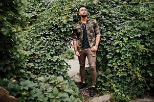 Awesome beautiful tall ararbian beard macho man in glasses and military jacket posed outdoor against arch of green leaves. photo