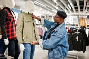 Stylish casual african american man at jeans jacket and black beret at clothes store looking on new jacket on mannequin. photo