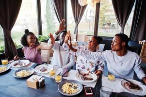 Happy african friends sitting, chatting in cafe and eat food. Group of black peoples meeting in restaurant and have dinner. Gives high five each other. photo