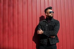 Fashion Arab man wear on black jeans jacket and sunglasses posed against red steel wall background. Stylish and fashionable arabian model guy. photo