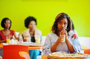 African woman with pizza sitting at restaurant and drink juice against dark skinned girls. photo