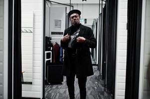 Stylish casual african american man at black beret and overcoat with waist bag at fitting room clothes store, looking on mirror. photo