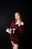 Young housewife in checkered dress with saucepan and kitchen spoon isolated on black background. photo