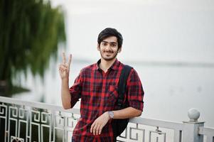 joven estudiante indio con camisa a cuadros y jeans con mochila posada en la ciudad nocturna contra el lago, muestra dos dedos. foto