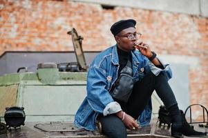 African american man in jeans jacket, beret and eyeglasses, smoking cigar and posed against btr military armored vehicle. photo