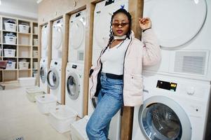 alegre mujer afroamericana con gafas de sol cerca de la lavadora en la lavandería de autoservicio. foto