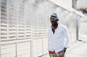 Stylish african american man in white shirt and colored pants with hat and glasses posed outdoor. Black fashionable model boy. photo