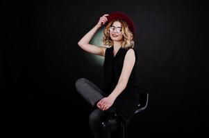 Studio portrait of blonde girl in black wear, red hat and glasses against dark background. photo