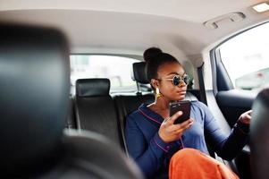 Rich business african woman on sunglasses sit at suv car with black leather seats. Mobile phone at hand. photo