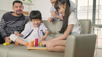 gelukkige aziatische mooie thaise gezinszorg, vader, moeder en kleine kinderen hebben plezier met het samen spelen met kleurrijke speelgoedblokken op de bank in de witte woonkamer, het vrijetijdsweekend en de huiselijke welzijnslevensstijl. video