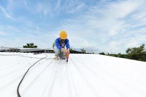 trabajador de la construcción en ropa de trabajo instalando nuevas herramientas para techos herramientas para techos, taladro eléctrico y uso en nuevos techos de madera con láminas de metal. foto