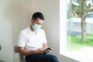 Asian man wearing mask and hold mobile phone text or message. Businessman use modern smartphone to browse web, read e-book, trade stocks. photo