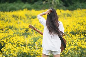 mujer joven asiática tocando la guitarra y cantando música en el parque, mujer asiática tocando la guitarra en el jardín de flores amarillas foto