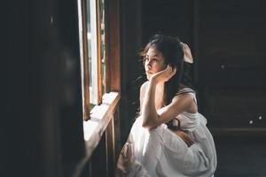 triste mujer asiática con vestido blanco que sufre depresión insomnio despierta y se sienta sola en la cama en un dormitorio antiguo. acoso sexual y violencia contra la mujer. foto