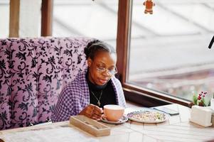 African woman in checkered violet cape and eyeglasses posed at cafe, sitting by table with dessert and cup of coffee. photo