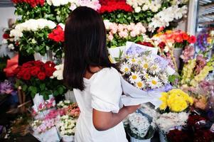 Beautiful african american girl in tender white dress with bouquet flowers in hands standing against floral background in flower shop.Black female florist. photo