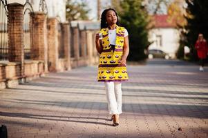 Stylish african american women in yellow jacket posed on street with hot drink in disposable paper cup. photo