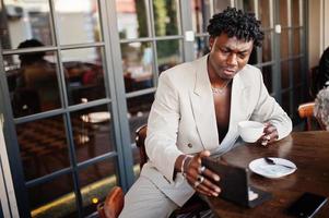 Stylish afro man in beige old school suit sitting on cafe with cup of coffee. Fashionable young African male in casual jacket on bare torso. photo