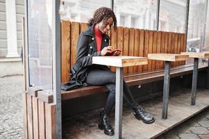 Portrait of a curly haired african woman wearing fashionable black coat and red turtleneck sitting with mobile phone at hands. photo