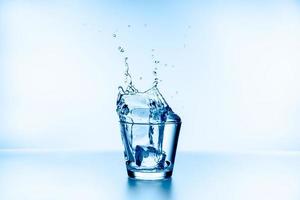 Hielo que cayó en el vaso con salpicaduras de agua de vidrio aislado sobre fondo azul. foto