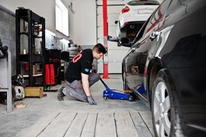 Car repair and maintenance theme. Mechanic in uniform working in auto service, lifted with hydraulic floor jack for repairing. photo