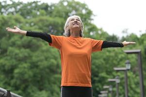 una mujer asiática jubilada con una camiseta naranja hace ejercicio al aire libre, corre, levanta pesas, se estira. foto