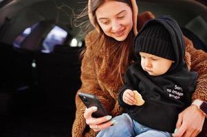 joven madre e hijo sentados en el maletero de un coche y mirando el teléfono móvil. concepto de conducción segura. foto