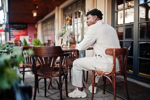 Stylish afro man in beige old school suit sitting on cafe with mobile phone. Fashionable young African male in casual jacket on bare torso. photo