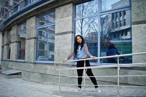 chica afroamericana hipster con camisa de jeans con mangas de leopardo posando en la calle contra un edificio de oficinas moderno con ventanas azules.