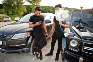 Two asian brothers man wear on all black posed near suv cars and smoke hookah. photo