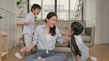 la joven madre asiática tailandesa se sienta en el piso de la sala de estar, medita y practica yoga para la salud y el bienestar, los niños juegan, caóticos y traviesos, feliz estilo de vida doméstico en el fin de semana familiar. video