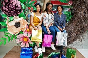 Beautiful three well-dressed afro american girls with colored shopping bags sitting on spring decoration photo zone in mall.