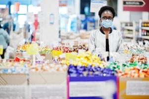 African woman wearing disposable medical mask and gloves shopping in supermarket during coronavirus pandemia outbreak. Black female choose candies at epidemic time. photo