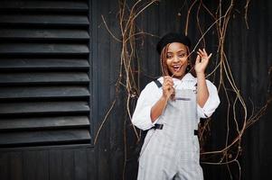 African american woman in overalls and beret posed outdoor against wooden background. photo