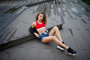 Chica modelo rizada sexy en top rojo, pantalones cortos de mezclilla, chaqueta de cuero y zapatillas de deporte posadas en el suelo. foto