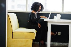 Fashionable young beautiful african american business woman with afro hairstyle wear in elegant black, sitting and working at laptop. photo