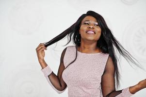 Fashionable african american model woman in pink brilliant evening dress posed against white decorative wall. photo