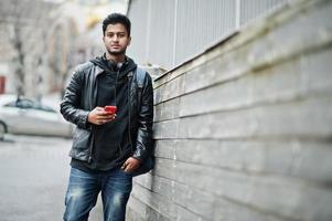 hombre asiático elegante e informal con chaqueta de cuero negro, auriculares con teléfono móvil rojo en las manos posado en la calle. foto