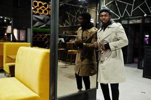 Handsome african american man posing  inside night club in black hat and beige coat. photo