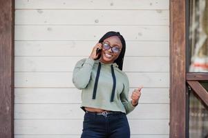 City portrait of positive young dark skinned female wearing green hoody and eyeglasses, speak on mobile phone. photo