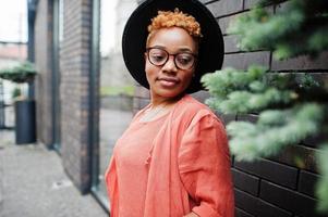 African red haired woman wear eyeglasses, orange dress and black hat posing outdoor. photo