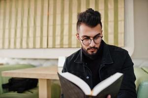 Arab man wear on black jeans jacket and eyeglasses sitting in cafe, read book. Stylish and fashionable arabian model guy. photo