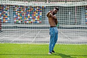 Handsome sexy african american muscular bare torso man at jeans overalls posed at green grass agasinst football gates of stadium field. Fashionable black man portrait. photo