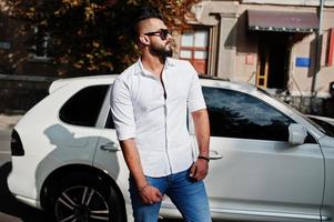 Elegante modelo de hombre árabe alto con camisa blanca, jeans y gafas de sol en la calle de la ciudad. chico árabe rico y atractivo con barba contra un auto todoterreno blanco. foto