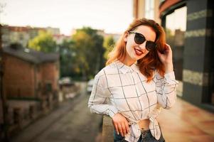 Attractive redhaired woman in sunglasses, wear on white blouse posing at street against modern building. photo