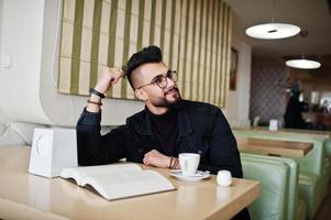 Arab man wear on black jeans jacket and eyeglasses sitting in cafe, read book and drink coffee. Stylish and fashionable arabian model guy. photo