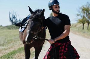 El hombre árabe de barba alta usa casco negro con caballo árabe. foto